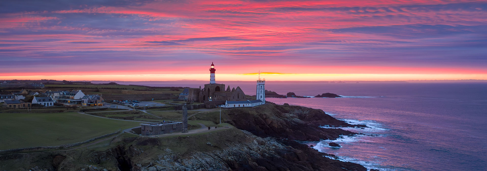 Superbe coucher de soleil sur la Pointe Saint-Mathieu dans les tons roses. L'ambiance est paisible, la mer est calme. Le phare et les commerces sont éclairés.