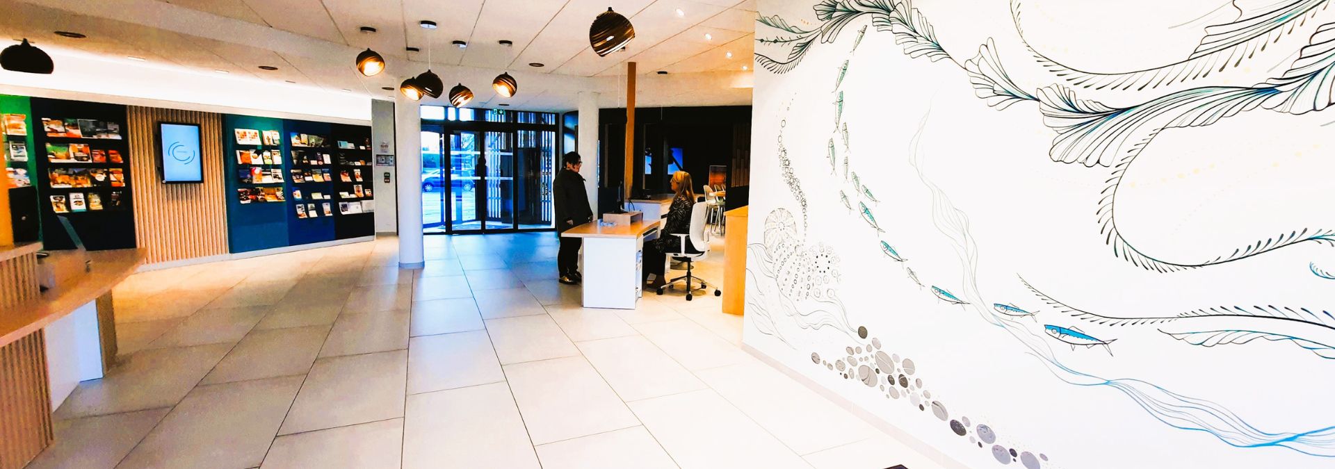 Hall moderne et lumineux de l'Office de Tourisme de Fouesnant-les Glénan avec comptoir d’accueil, présentoirs et fresque murale illustrée.