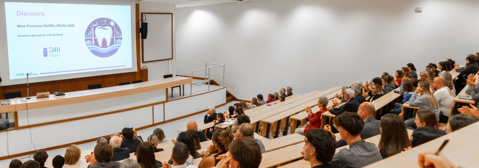 Conférence dans un amphithéâtre universitaire avec un public attentif et un écran de présentation.